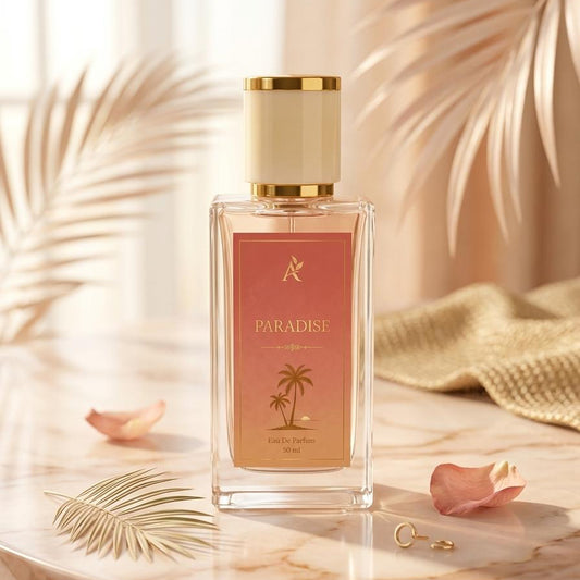 A rectangular glass perfume bottle labeled 'PARADISE' with a pink label and gold cap, styled on a marble surface with palm leaves and soft warm background elements.